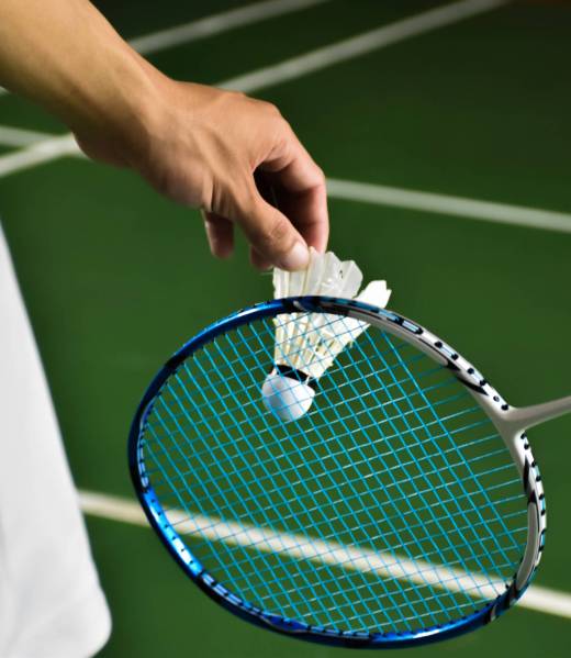 Badminton player is holding white badminton shuttlecock and badminton racket in front of the net before serving it over the net to another side of badminton court. Selective focus on white shuttlecock